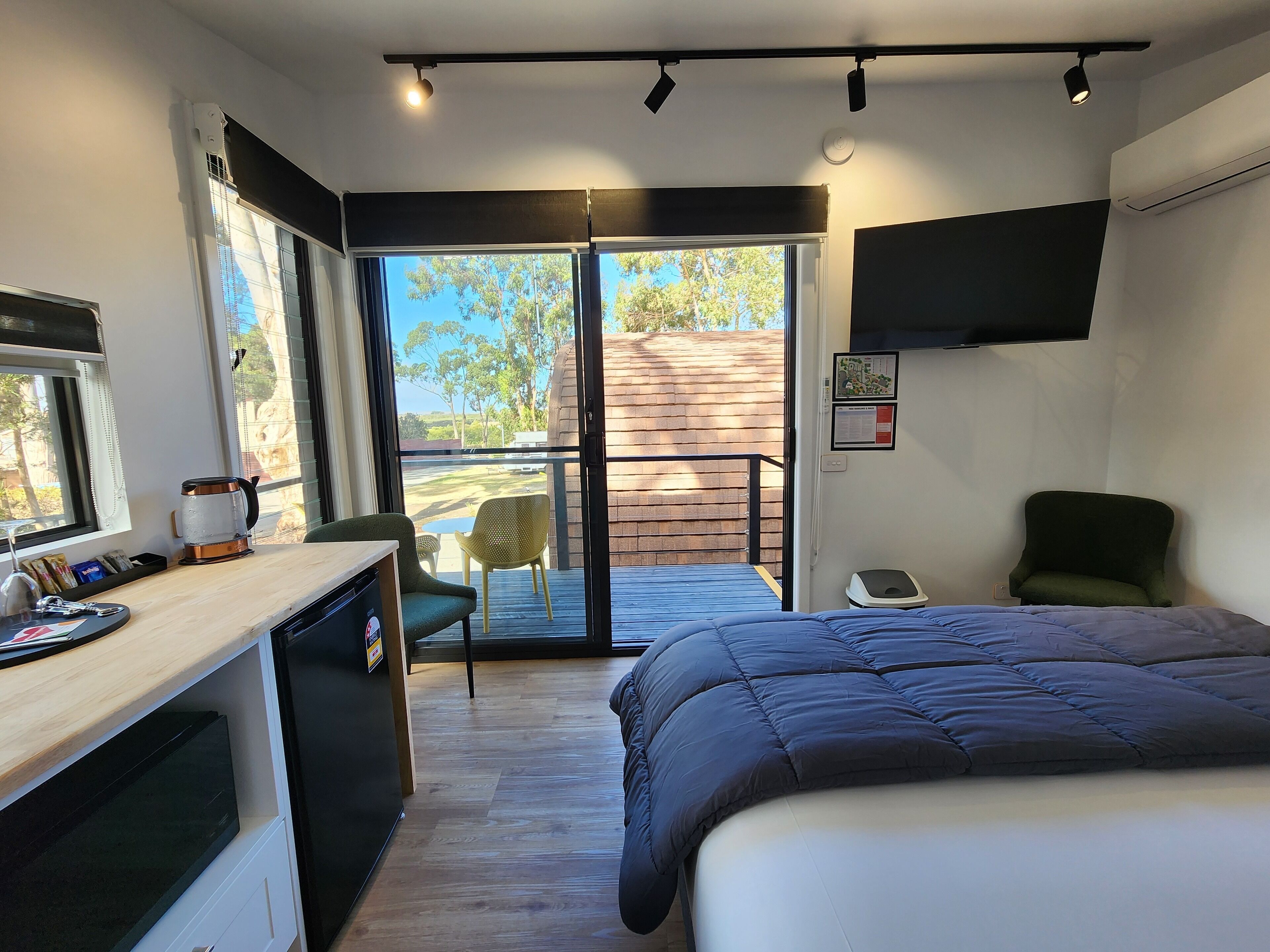 A modern bedroom with a bed, a desk with a kettle, a chair, and a TV mounted on the wall.