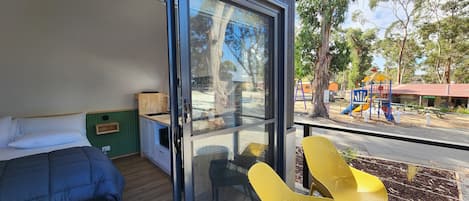 A modern room with a bed, a small kitchenette, and a sliding glass door leading to an outdoor area with yellow chairs and a playground.