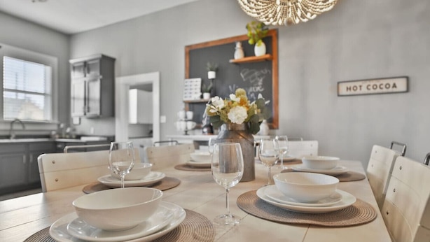 Comedor con una mesa dispuesta para cuatro, que incluye cuencos, platos y copas de vino. Un jarrón con flores está sobre la mesa, y un letrero en la pared dice "HOT COCOA".