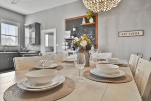 A dining area with a table set for four, including bowls, plates, and wine glasses. A vase with flowers is on the table, and a sign on the wall reads "HOT COCOA". - Sparkled Seahorse (Galveston)