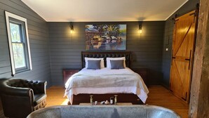 A bedroom with a large bathtub, a bed with white linens, a dark leather chair, and a painting of a lakeside scene. - Murray River Spa Retreat (Echuca)