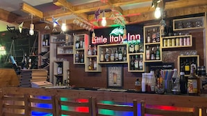 Zona de bar con vigas de madera, estantes con botellas de licores y un letrero de neón que dice "Little Italy Inn".