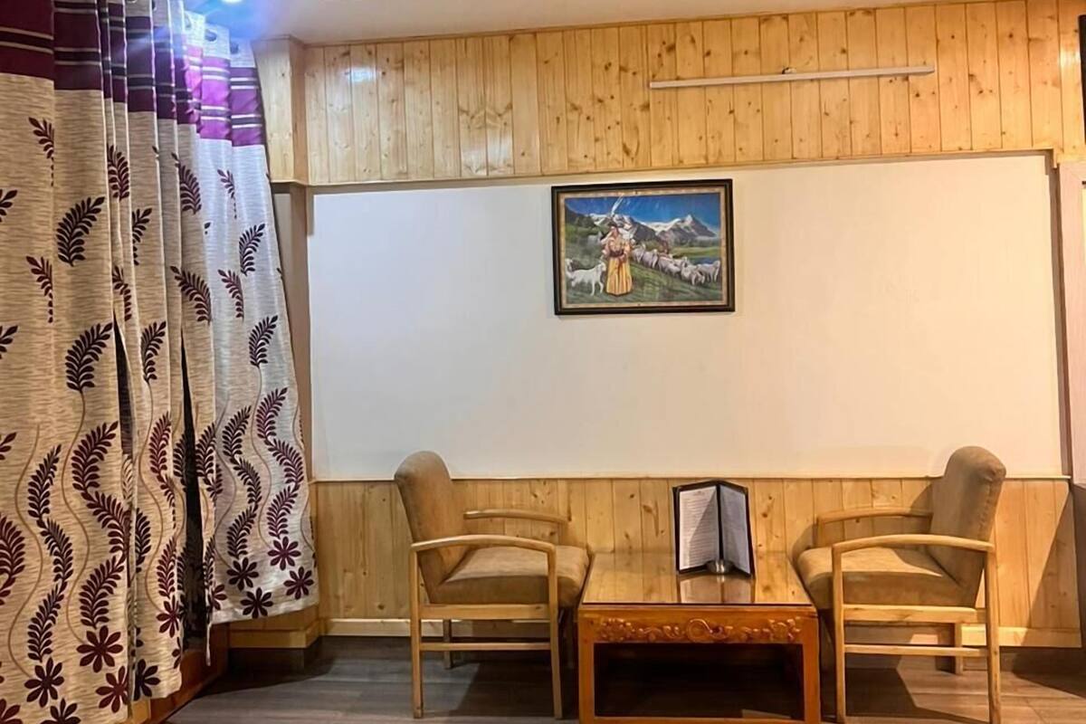 A room with a bed, two chairs, a small table, wooden panel walls, and a painting on the wall.