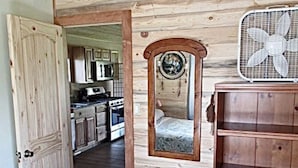 A rustic interior with wooden walls, a kitchen visible through an open door, a mirror, and a fan. - Dreamcatcher Bungalow 20min to W.Glacier, between Glacier&Airport (Columbia Falls)