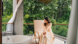 Een vrouw zit bij een badkuip op een balkon met uitzicht op weelderige begroeiing.
