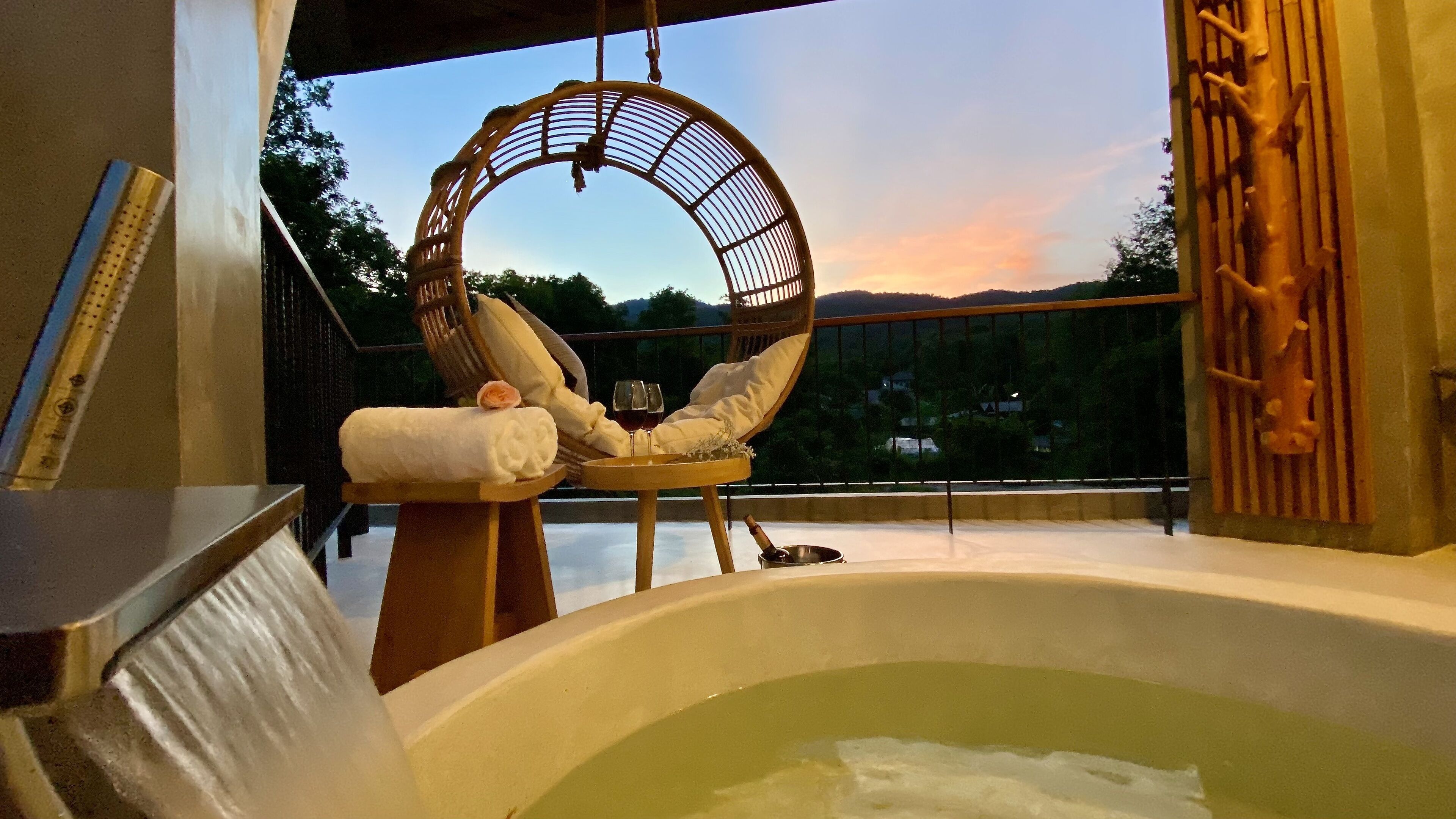 Un jacuzzi con una silla de mimbre, una toalla, una botella de champán y una copa de vino tinto en un balcón con vistas panorámicas.