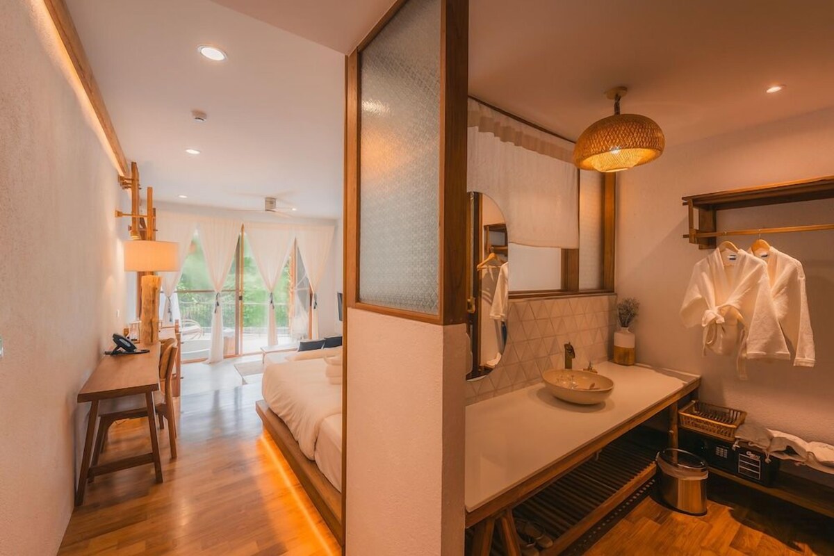 Une chambre d’hôtel avec un lit, un bureau, une salle de bain avec un lavabo et un miroir, et une vue sur les arbres à l’extérieur.