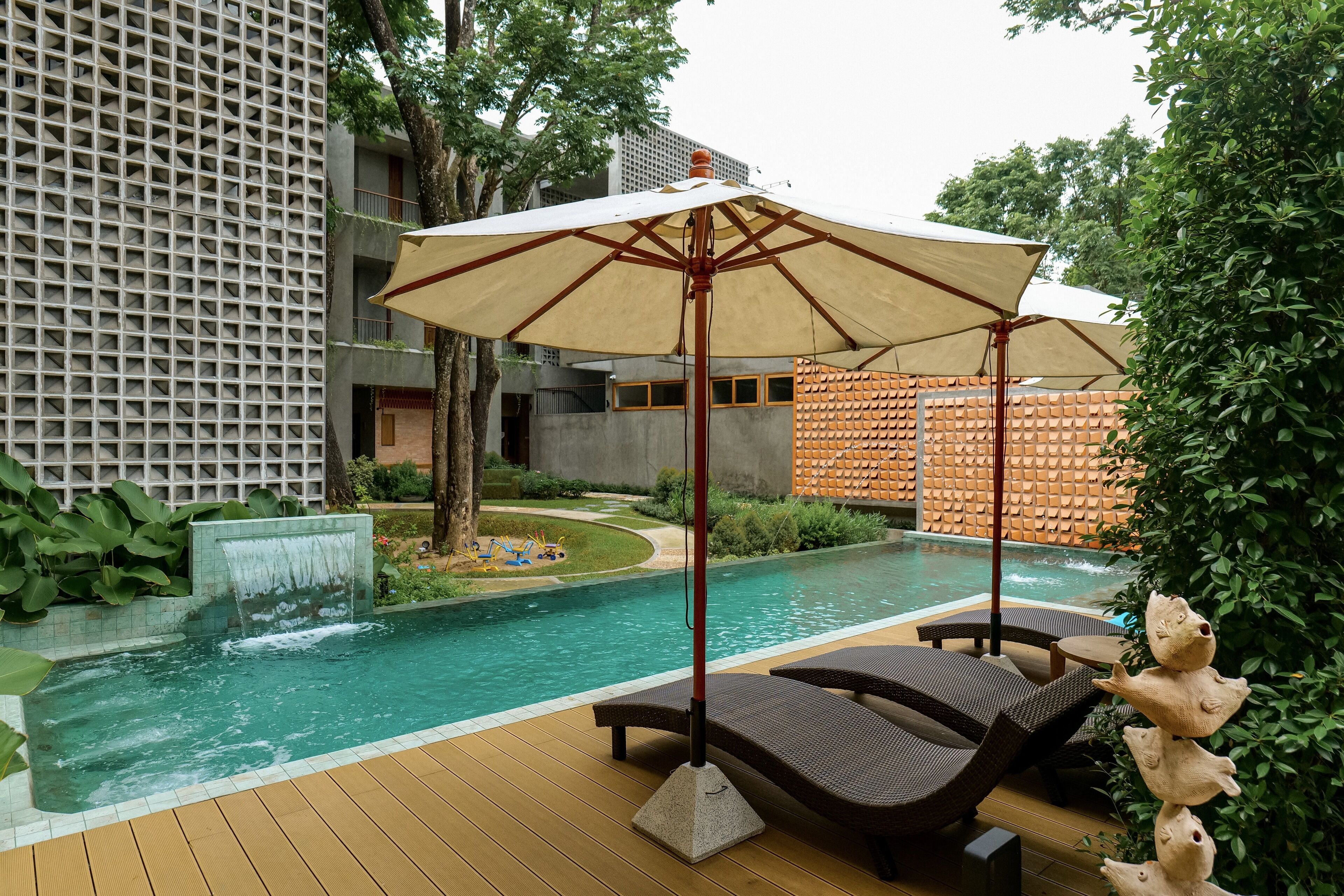 Zona de piscina con sillones de descanso y un toldo, rodeada de follaje y un edificio moderno.