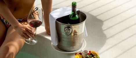 Una persona en traje de baño sentada a una mesa con una botella de champán en un cubo, una copa de vino tinto y un plato de aperitivos.