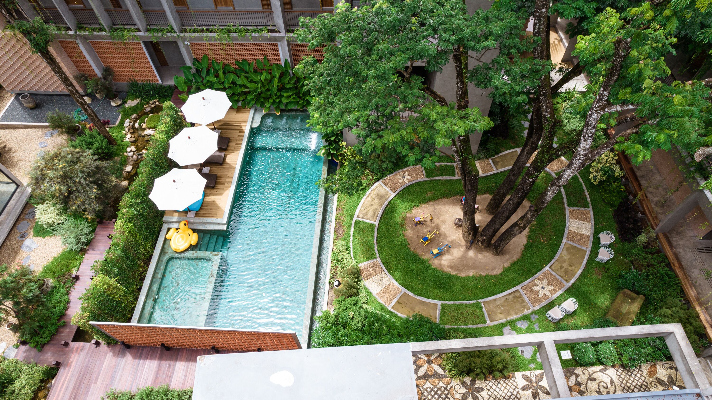 An aerial view of a rooftop pool area with a rectangular pool, surrounded by lush greenery, trees, and outdoor seating.
