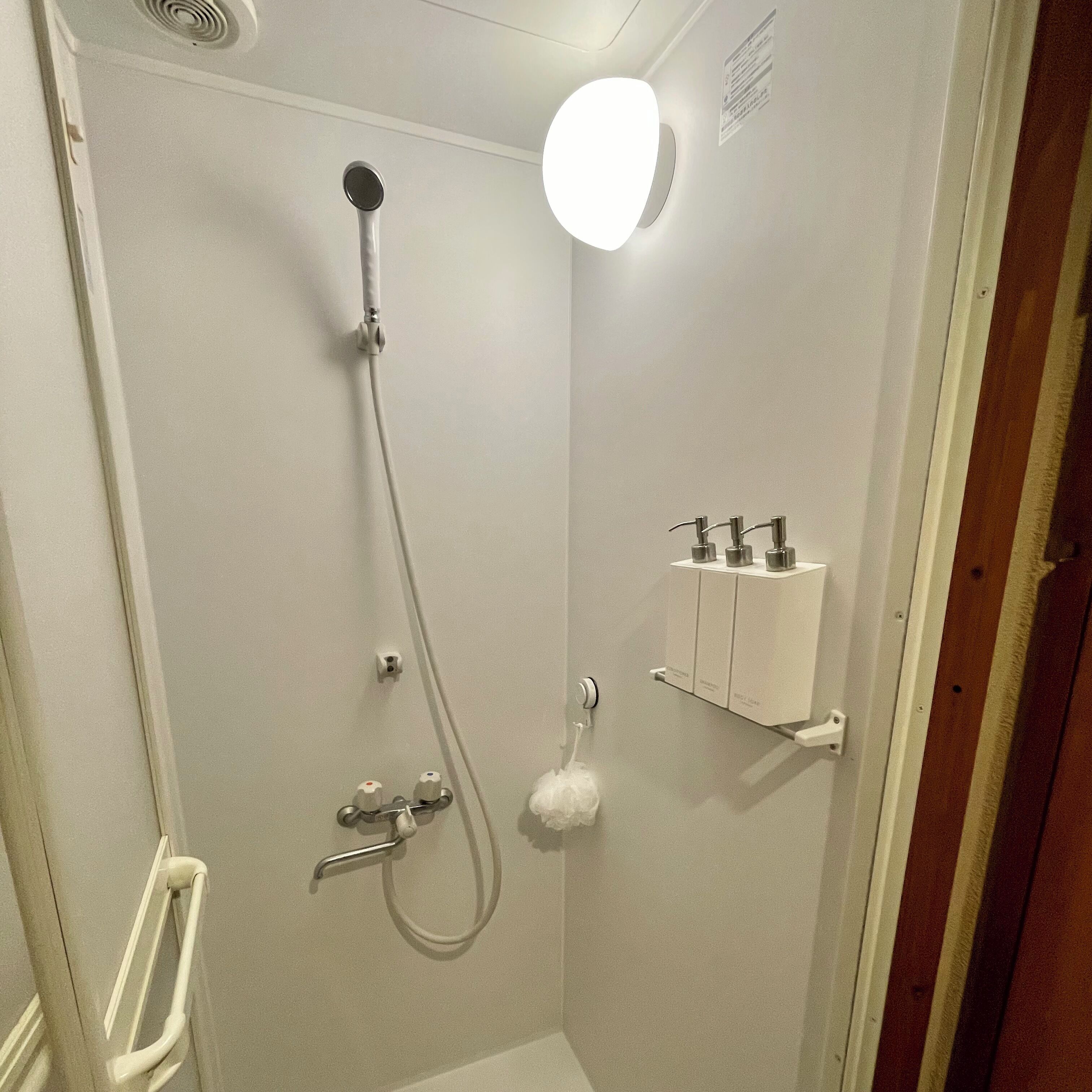 A small, enclosed shower area with a handheld showerhead, a wall-mounted light, and a soap dispenser.