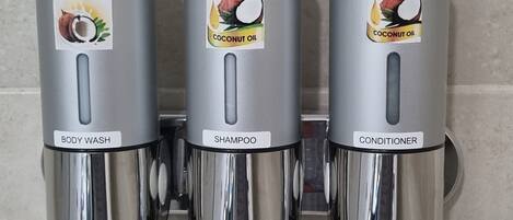 Tres dispensadores metálicos etiquetados con 'COCO' y diferentes tipos de producto: jabón corporal, champú y acondicionador.
