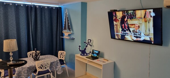 A room with a TV mounted on the wall displaying a show, a table with chairs, a lamp, and a decorative sailboat.