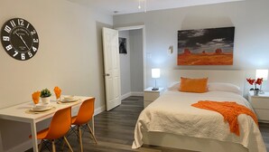 A hotel room with a bed, a desk with chairs, a clock, and a painting on the wall.