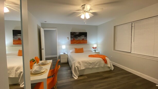 A hotel room with two beds, a dining area with a table and chairs, a ceiling fan, and artwork on the walls.
