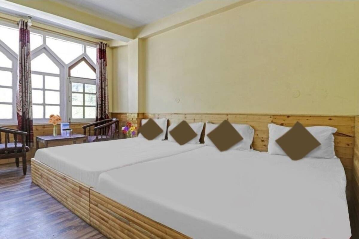 A room with two beds, a wooden floor, a window with curtains, and a small table with a vase.
