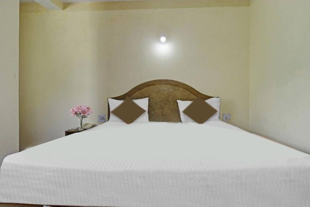 A neatly made bed with a wooden headboard and decorative pillows, a small bedside table with a vase of flowers, and a single light fixture on the ceiling.
