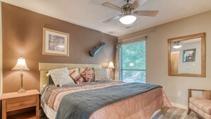 Une chambre à coucher comprenant un lit, des tables de chevet, un ventilateur de plafond, une fenêtre avec des stores et un miroir au mur.