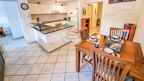 A modern kitchen with white cabinets, a black countertop, and a dining area with wooden chairs and a table set for two. - ZEN CITY MODERN - 1 BR City Apartment (Larrakeyah)