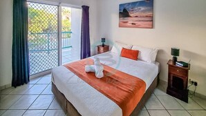 A hotel room with a bed, a nightstand, a lamp, a painting on the wall, and a view through a glass door. - ZEN CITY MODERN - 1 BR City Apartment (Larrakeyah)