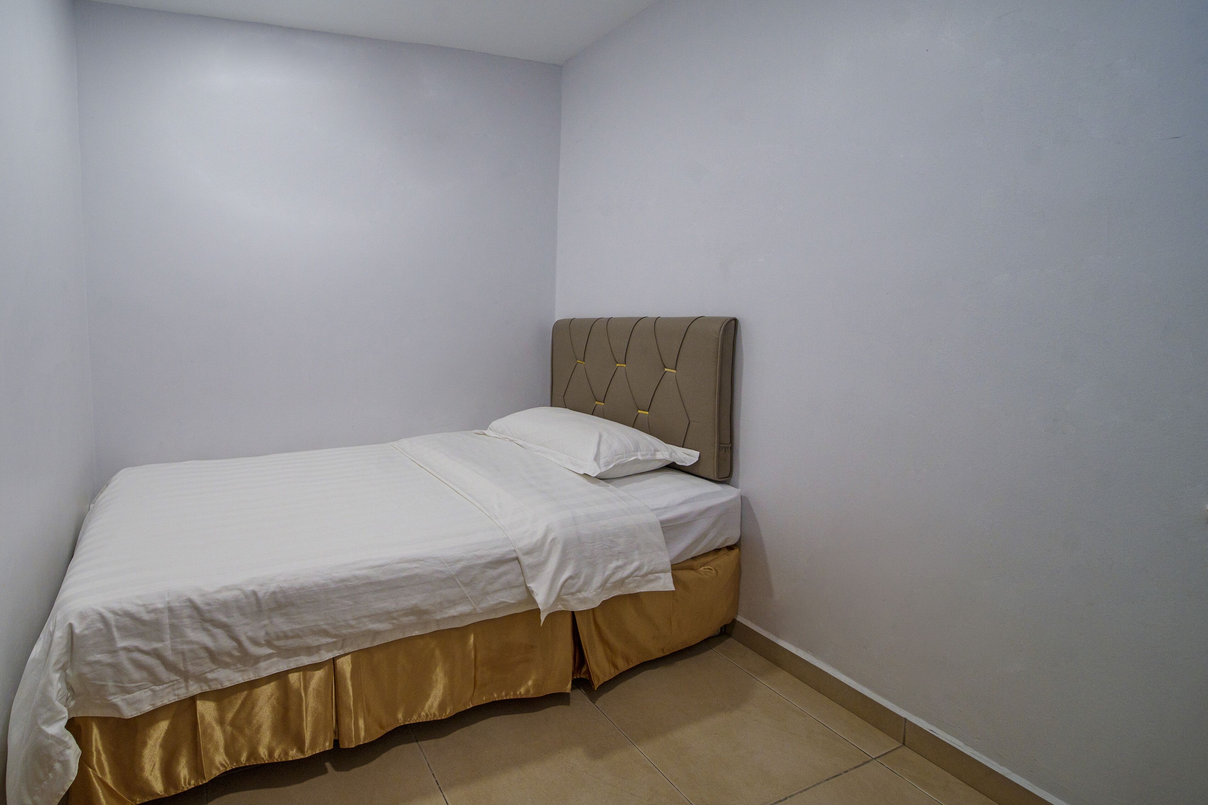 A small, neatly made bed in a corner room with plain walls and a simple headboard.