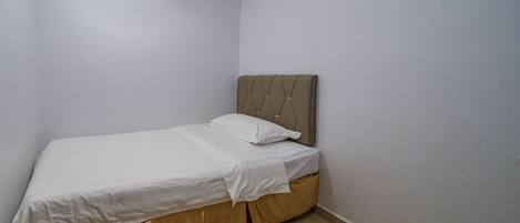 A small, neatly made bed in a corner room with plain walls and a simple headboard.