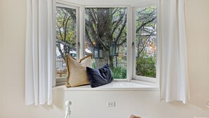 A bay window with white curtains, a cushion, and a view of trees and a street.