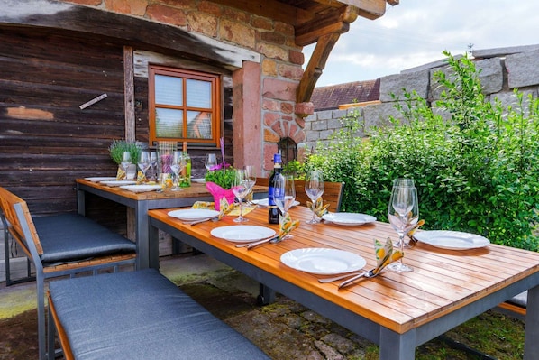 Un área de comedor al aire libre con mesas y sillas de madera, un muro de piedra y follaje.