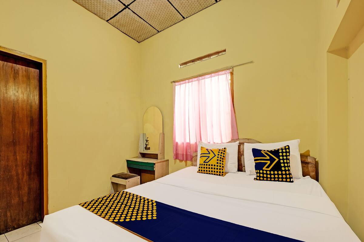 Um quarto com uma cama, uma mesa de cabeceira de madeira, um espelho, uma janela com cortinas e uma porta.