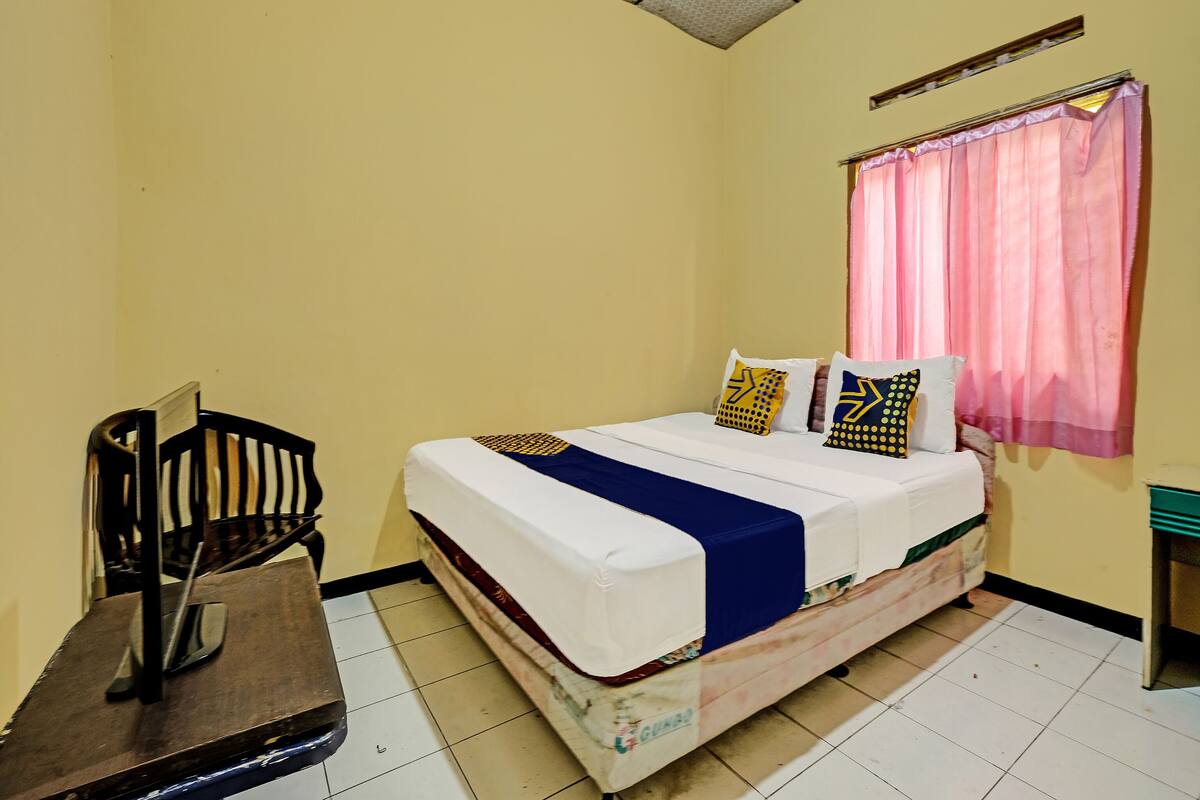 Quarto com cama, almofadas, uma cadeira, escrivaninha e janela com cortinas.