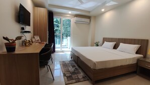 A modern hotel room with a large bed, a desk with a flat-screen TV, a potted plant, and a view of the outdoors through a sliding glass door. - Covie Gurgaon 42 (Gurugram)