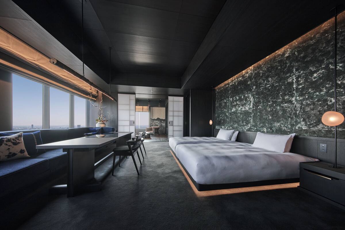 A modern bedroom with a large bed, a desk, and a sofa. There is a view of the city through large windows.