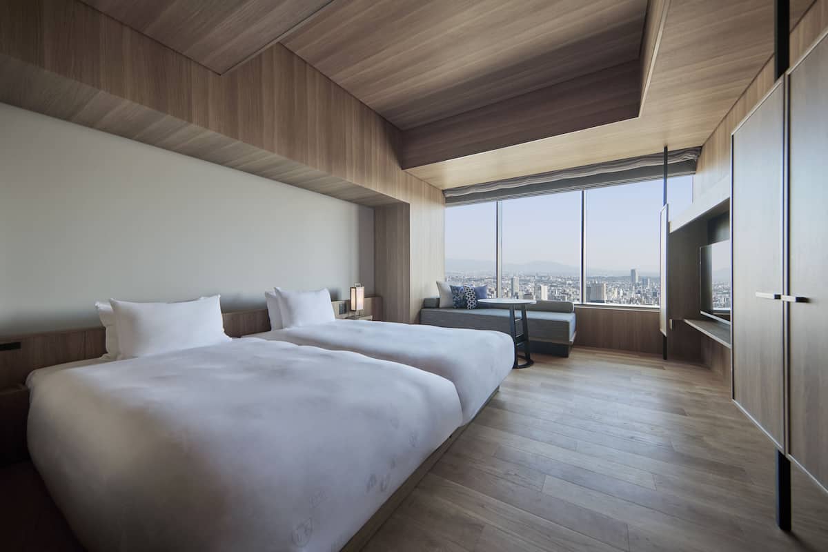 A modern hotel room with two large beds, a desk, and a large window offering a city view.