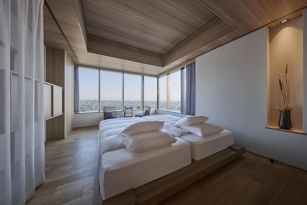 A modern bedroom with a large bed, wooden flooring, and a city view through large windows.