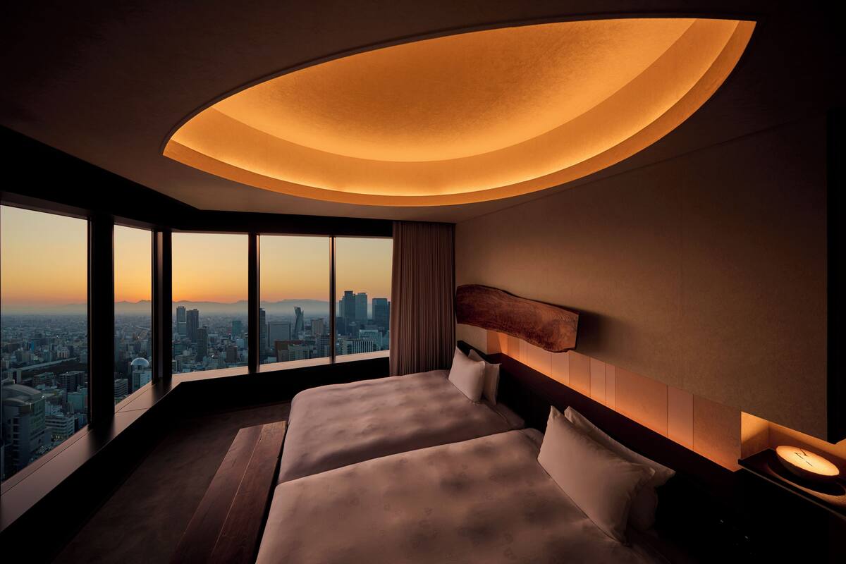 A hotel room with a city view, two beds, a curved ceiling light, and a nightstand with a clock.