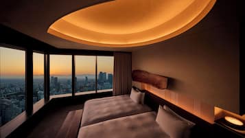 A hotel room with a city view, two beds, a curved ceiling light, and a nightstand with a clock.