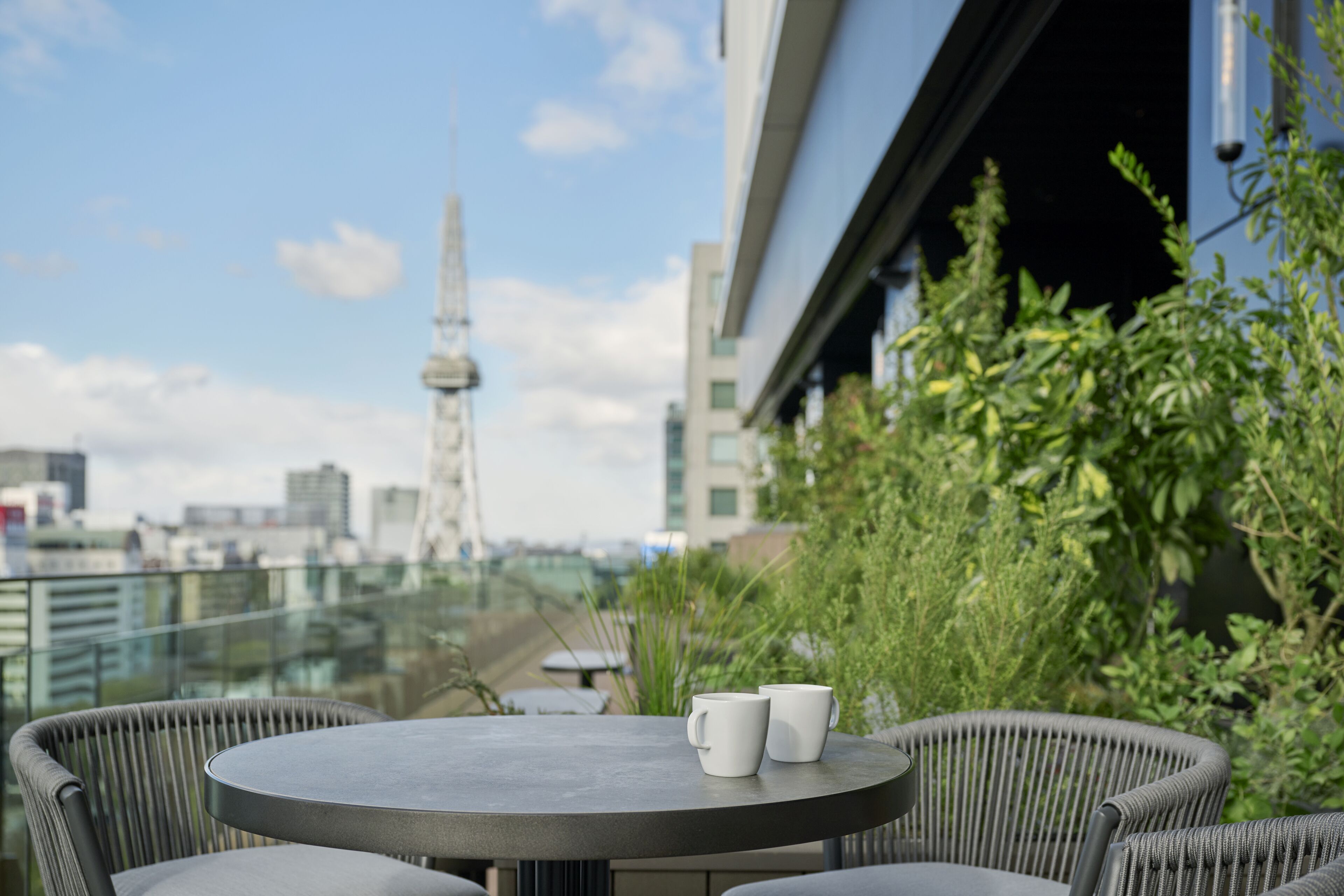 Una mesa redonda con dos tazas blancas en un balcón con vista a una ciudad, con edificios modernos y una torre.
