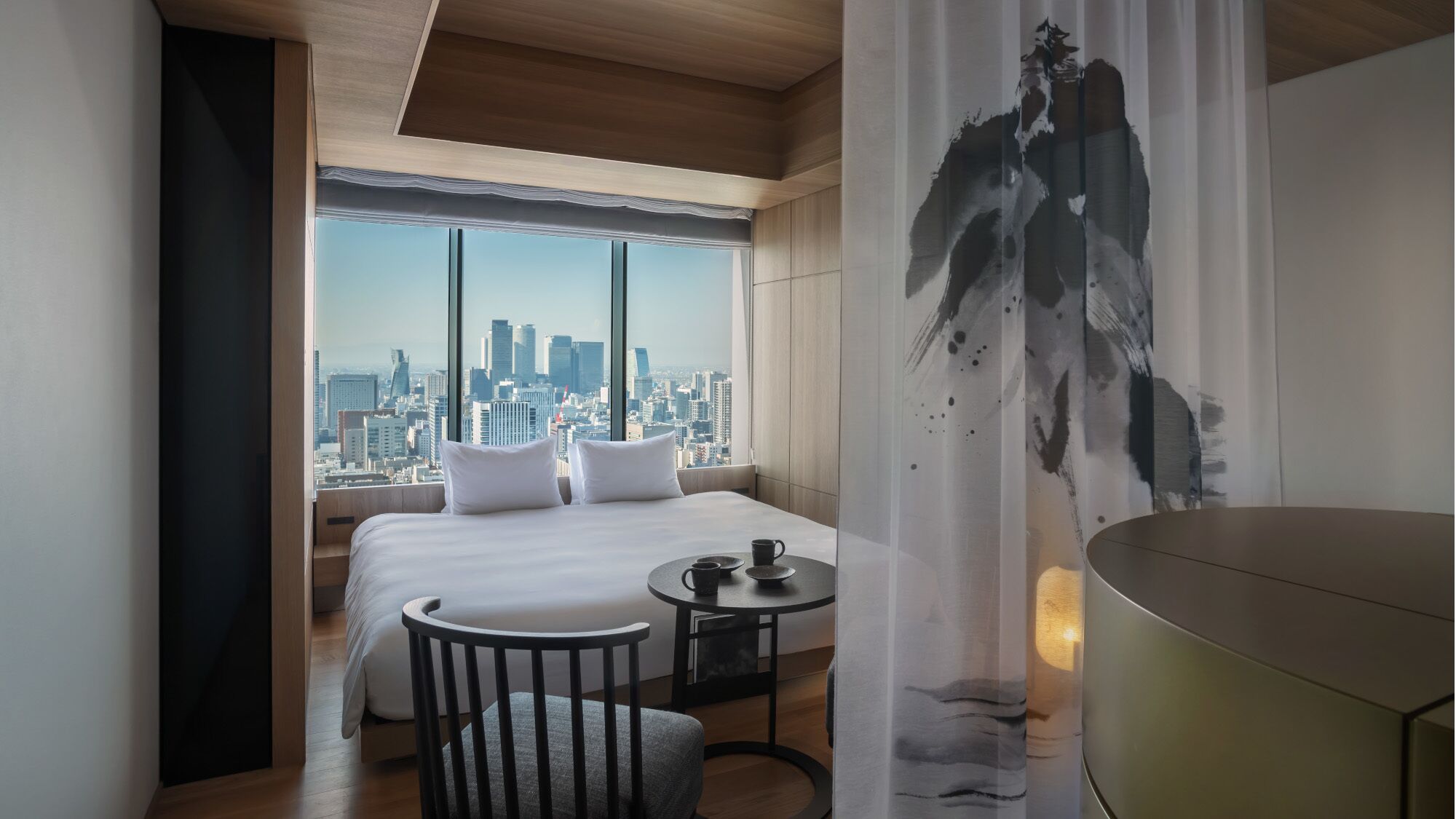 Camera d'albergo con un letto grande, un tavolino con una tazza, una sedia e vista sulla città attraverso ampie finestre.