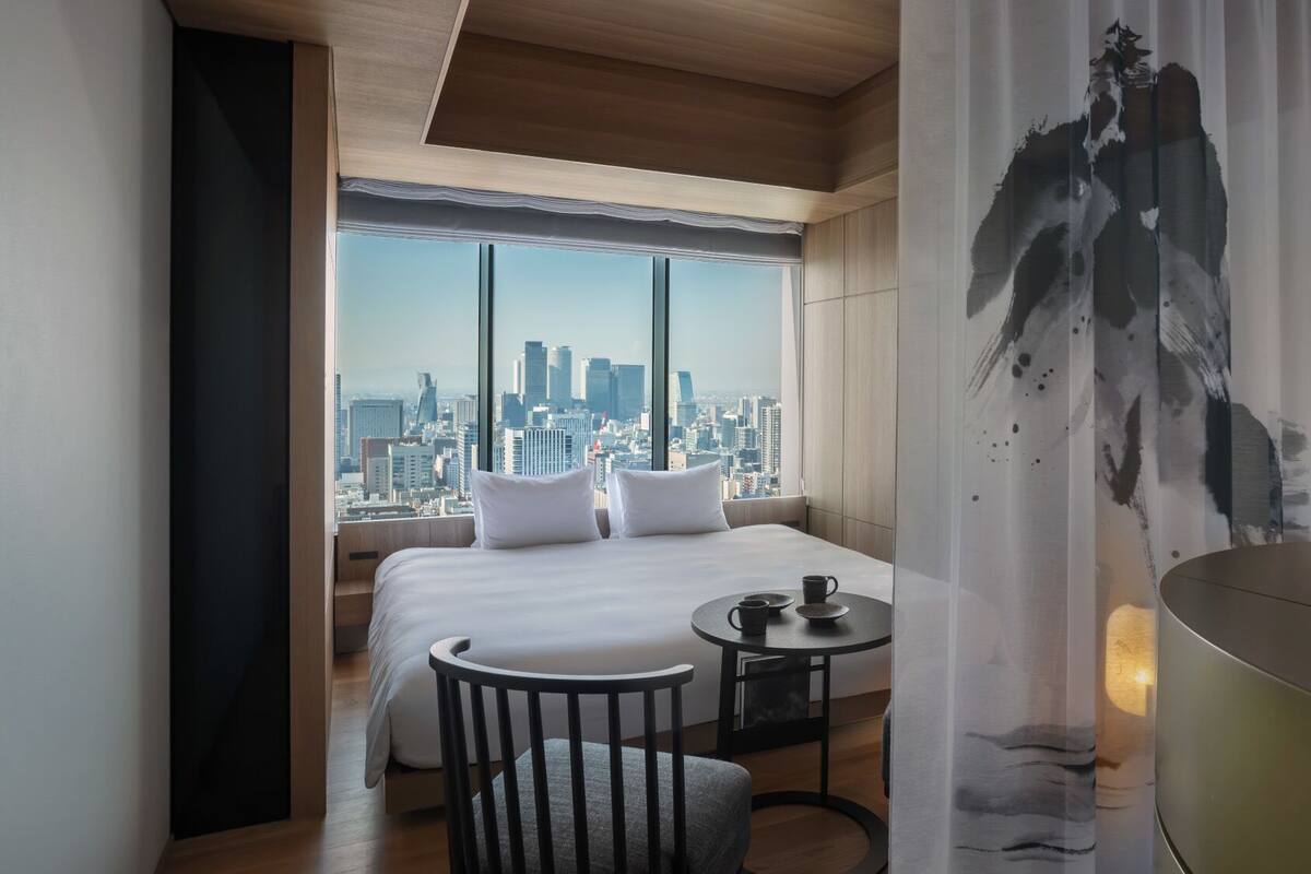 A hotel room with a large bed, a small table with a cup, a chair, and a view of a cityscape through large windows.