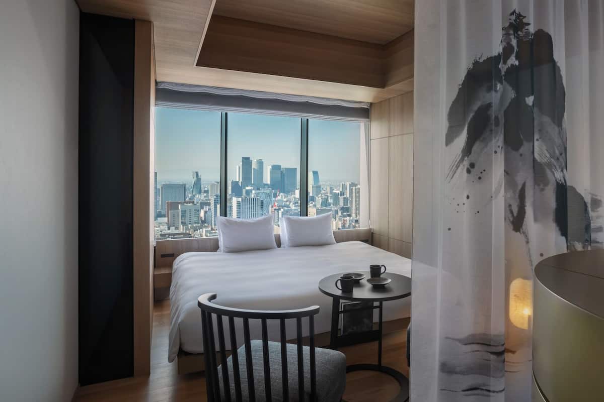 Une chambre d’hôtel avec un grand lit, une petite table avec une tasse, une chaise et une vue sur le paysage urbain à travers de grandes fenêtres.