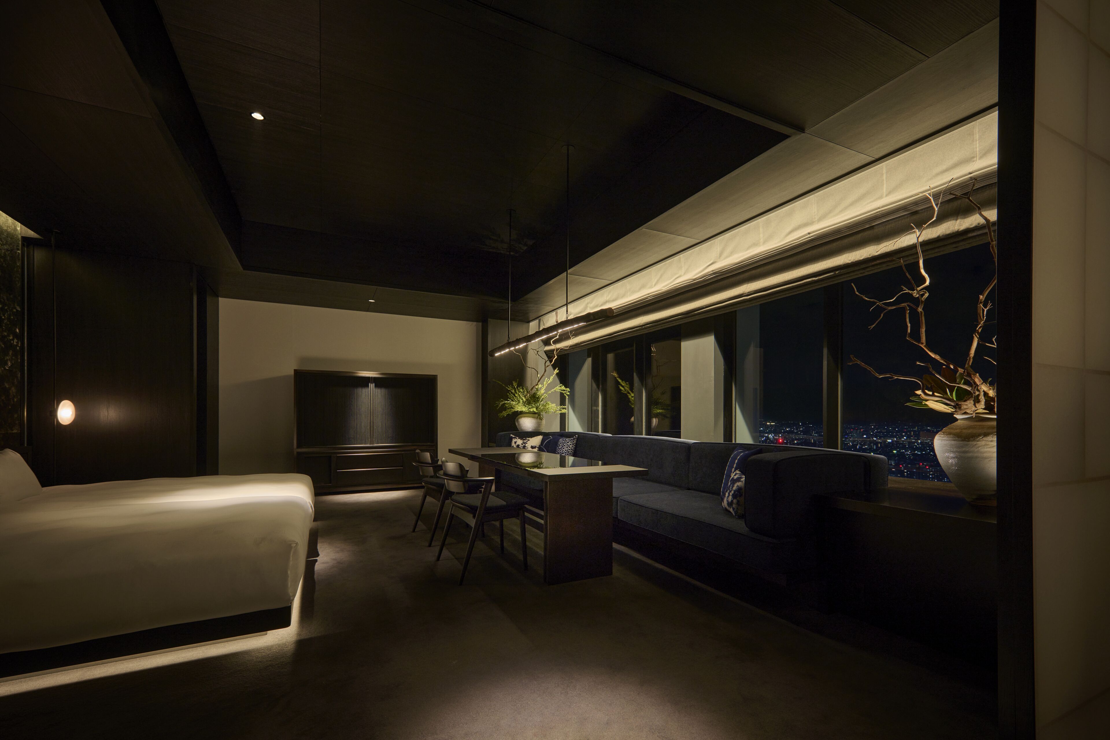 A modern hotel room with a sofa, a dining table, and a TV. The room has a large window with a view of the city at night.