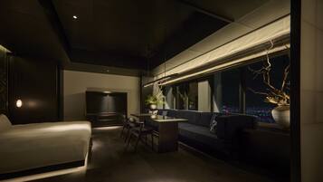 A modern hotel room with a sofa, a dining table, and a TV. The room has a large window with a view of the city at night.