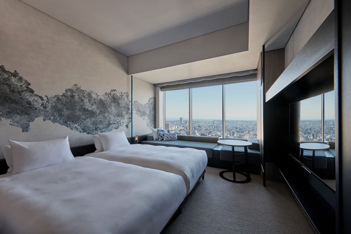 A modern hotel room with two beds, a large window with a city view, a round table, and a seating area with a view.