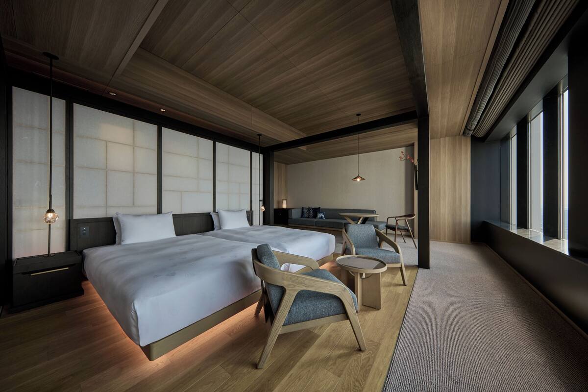 A modern hotel room with a large bed, two armchairs, a small table, and a view of the city through large windows.