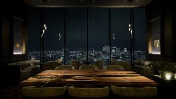 A modern dining area with a large wooden table and comfortable seating, overlooking a cityscape at night.