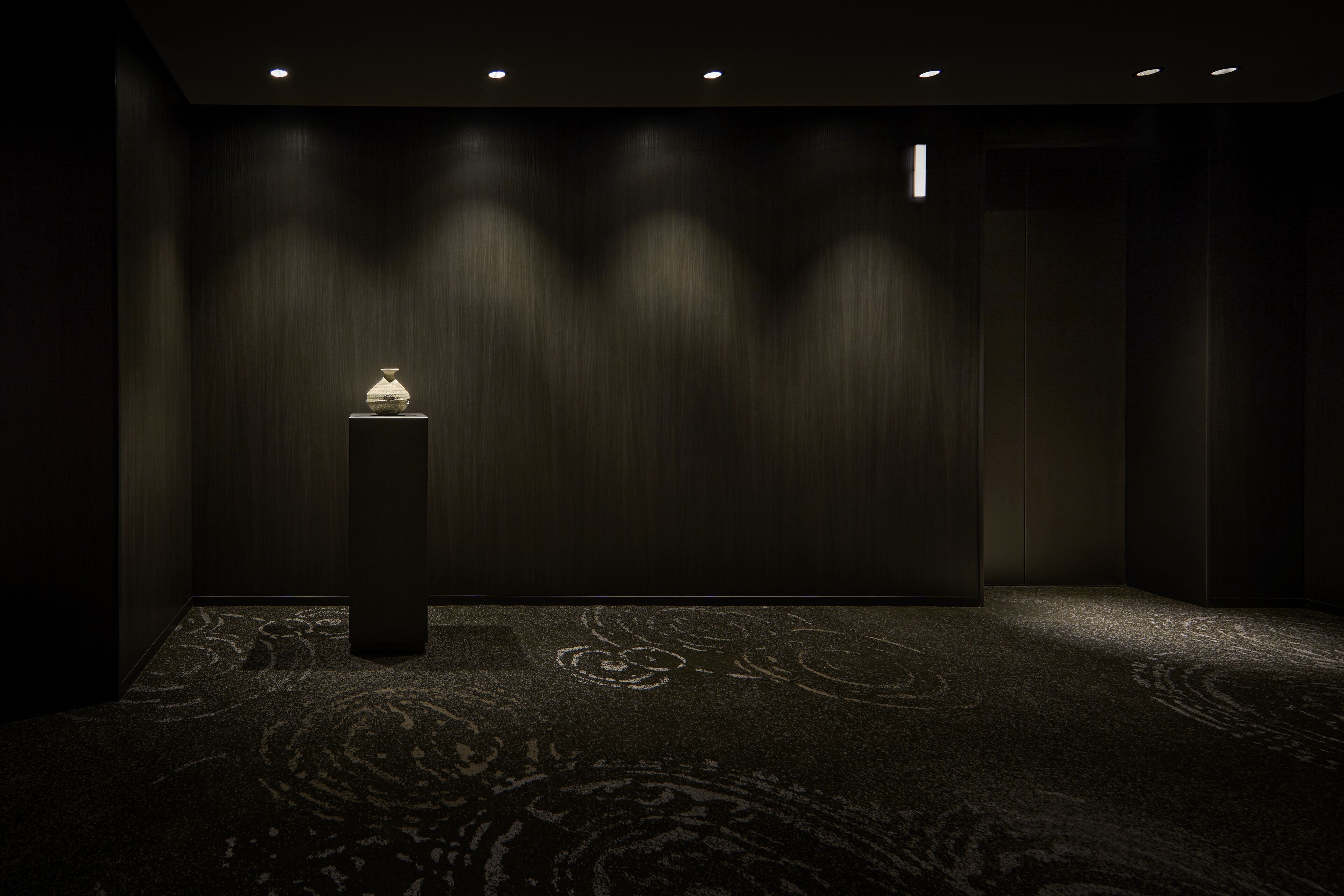 Une pièce faiblement éclairée, dotée d’un tapis à motifs, d’un haut pilier surmonté d’une lampe et d’un mur en bois sombre.