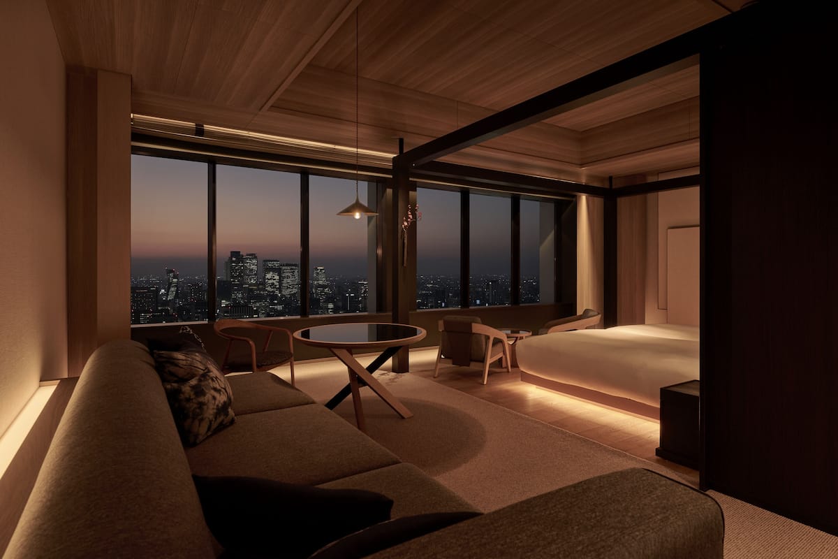 A modern hotel room with a large window offering a city view, a cozy seating area, and a minimalist design.