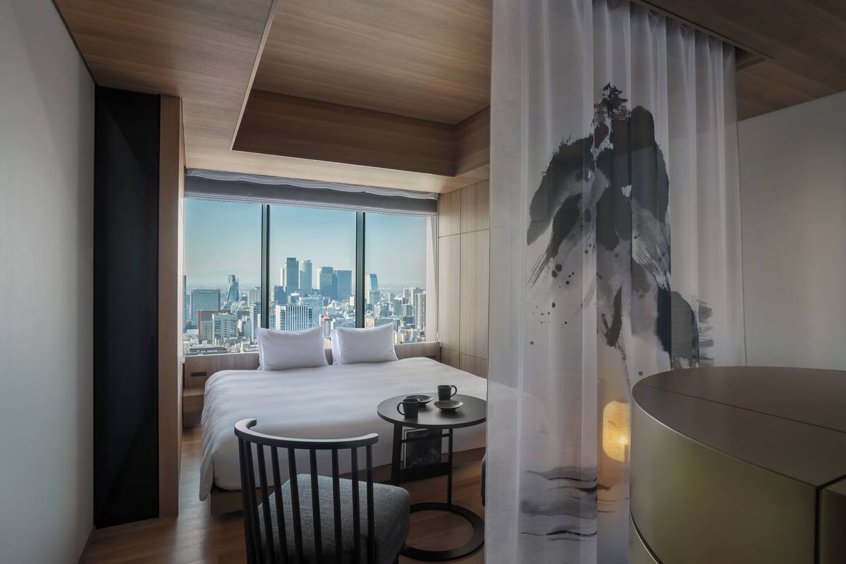 A hotel room with a large bed, a small table, a chair, and a view of a cityscape through a large window.