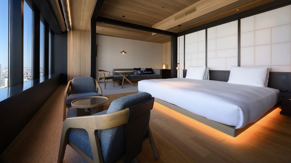 Una moderna camera d'albergo con un letto grande, una zona salotto con sedie, un tavolino e vista sulla città attraverso ampie finestre.
