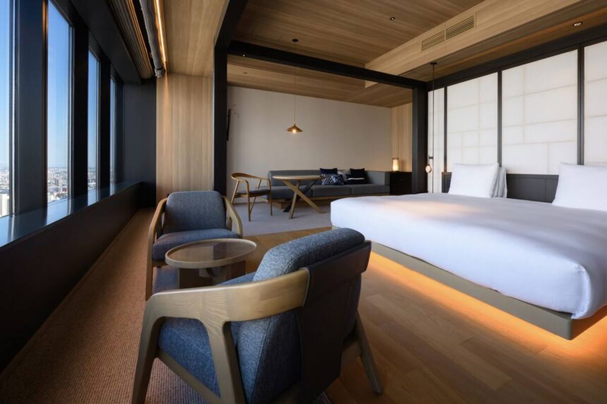 A modern hotel room with a large bed, a seating area with chairs, a small table, and a city view through large windows.
